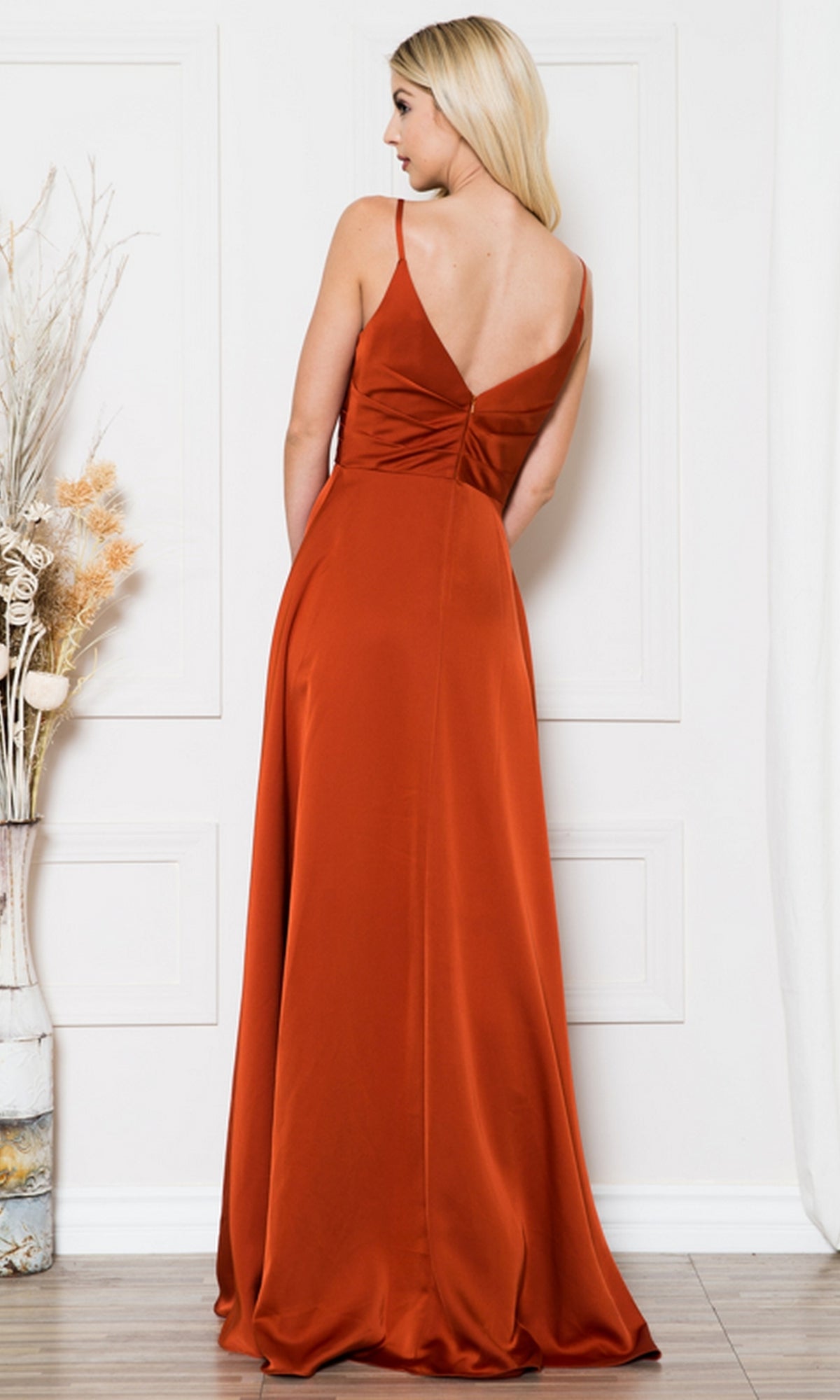 A woman wears the Long Satin A-Line Formal Prom Dress BZ012 in burnt orange, posing with her back to the camera in a white room decorated with dried plants.